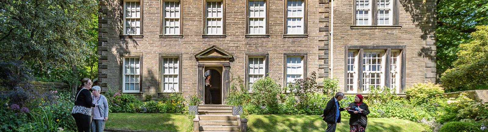 Photo of the front of the Bronte Parsonage Museum on a sunny day, with visitors in the front garden.