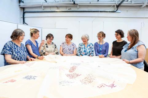 Photo of eight women grouped around a large piece of white fabric with sections of colour.