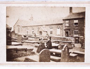 A black and white photo of a Victorian woman and man in a graveyard with a school house in the background.