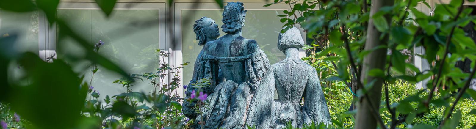 Photo of a statue of three women, seen through foliage.