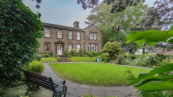 Photo of a green garden with an 18th-century house in the background and a bench in the foreground.