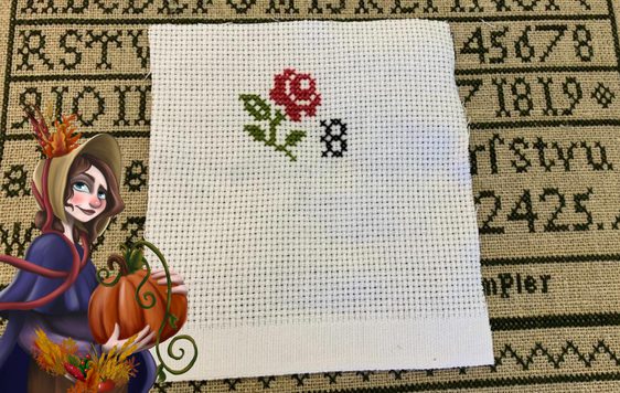 Photo of cross-stitching, illustration of a woman holing a pumpkin in bottom left corner.