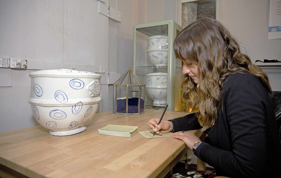 Photo of a young woman writing on a note card opposite a ceramic pot.