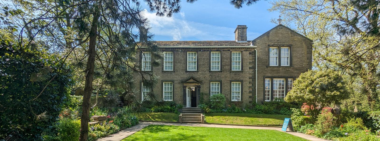 Photo of the Bronte Parsonage Museum on a sunny day, framed by trees and garden.