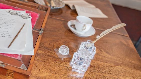 Photo of a quill in a glass ink pot on a table with assorted items.