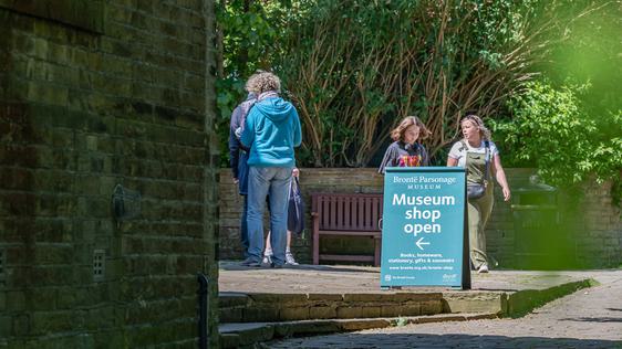Photo of people walking in the sunshine behind a sign point to a museum shop.
