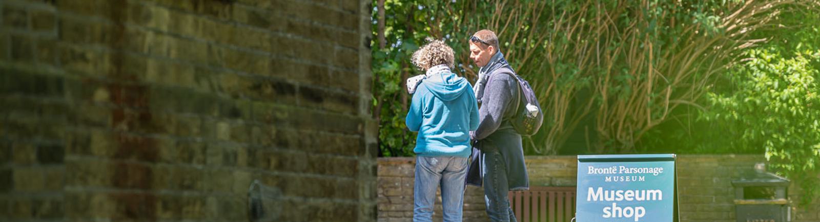 Photo of two people standing by a building, next to a sign reading 'museum shop open'.