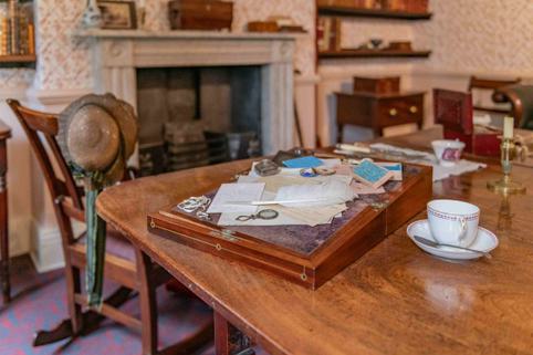 Photo of a writing desk with all its contents including papers and a quill on the dining table, next to a cup and saucer.