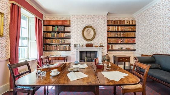 Photo of the Dining Room, with the dining table, sofa and bookshelves. A portrait of Charlotte hangs in the centre of the wall.