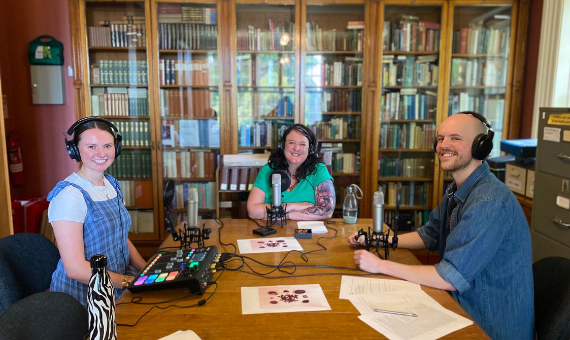 Three people sat round a table in a library with microphones.