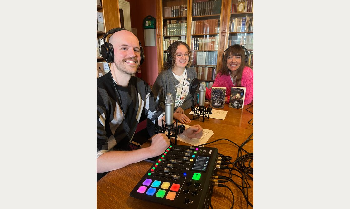 Three people wearing headphones, sat in a library with microphones on the table.