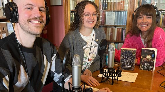 Three people wearing headphones, sat in a library with microphones on the table.
