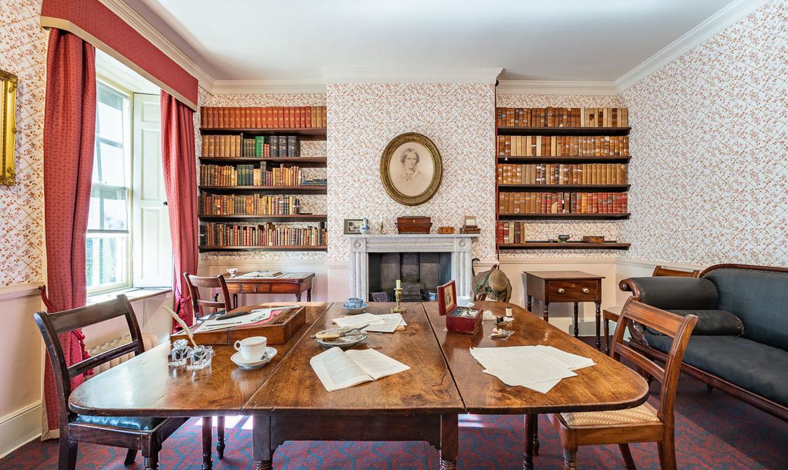 The dining room with the table and chairs in the centre of the room, fireplace at the back with bookshevles at either side. A green sofa on the right hand wall.