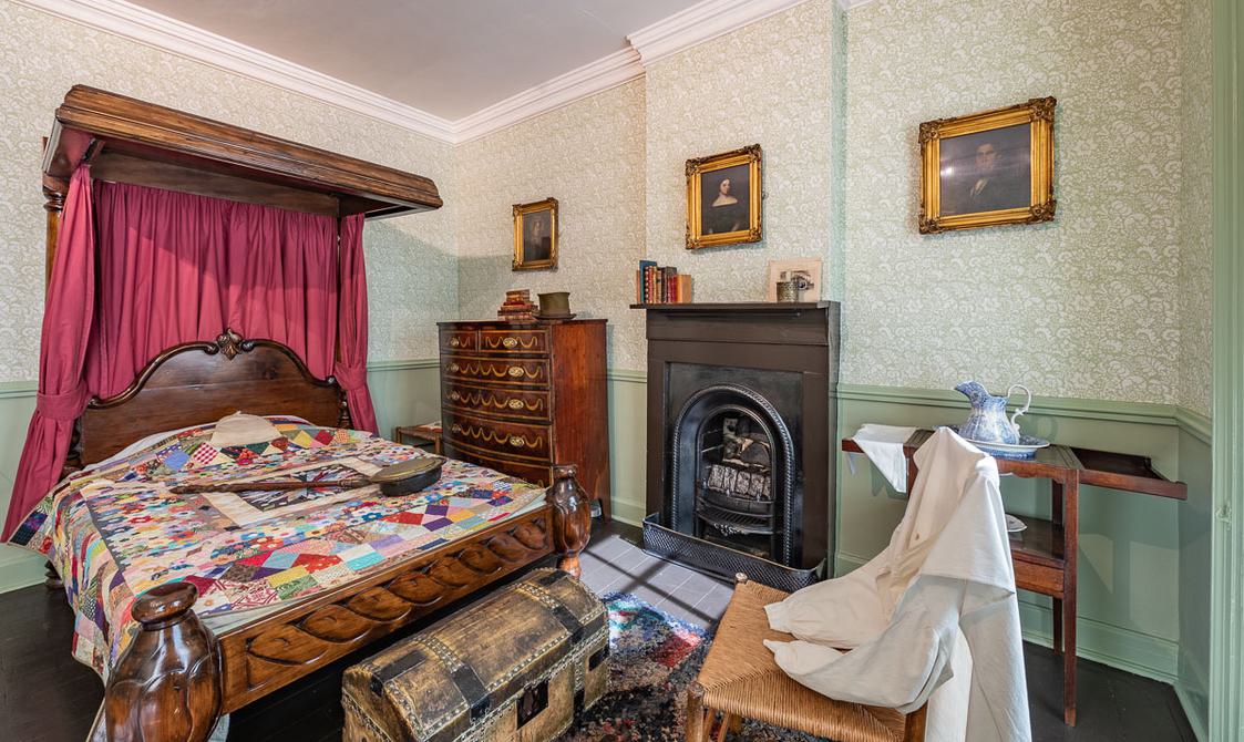 A bedroom with green wallpaper, a bed with a colourful quilt, portrait paintings by Branwell on the wall, a fireplace and chair with a white nightgown on.