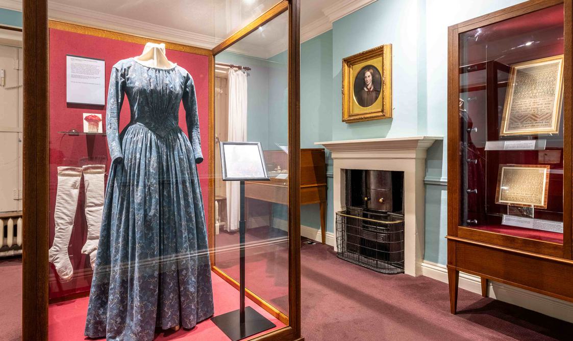 Room with teal walls, glass cases holding victorian clothing. Charlotte's portrait hangs above the fireplace.