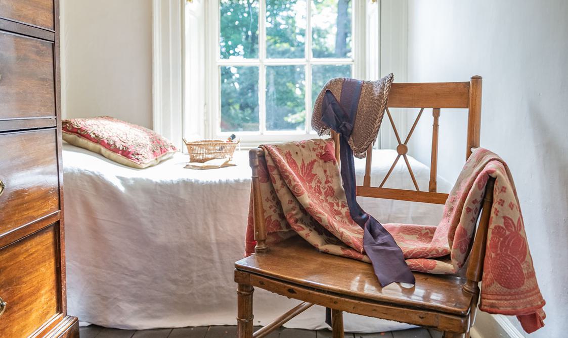 A small room containing a chair with a shawl and bonnet on, a small bed and window.
