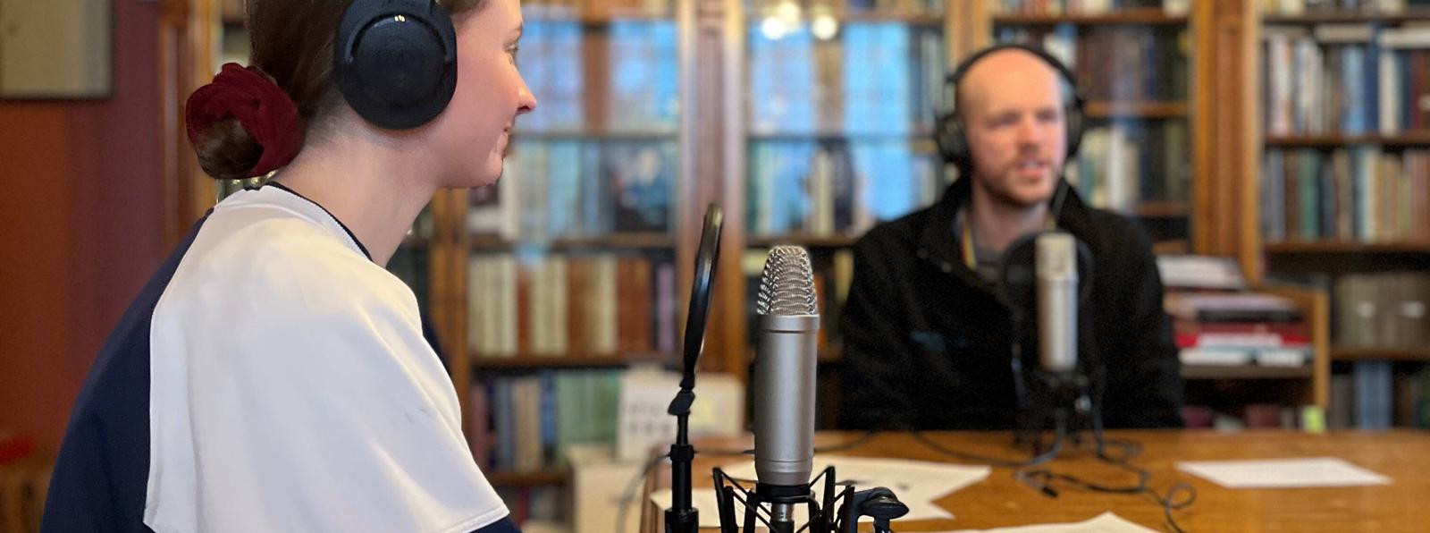 Photo of a young woman with headphones on in front of a microphone, and a young man in the background wearing headphones.