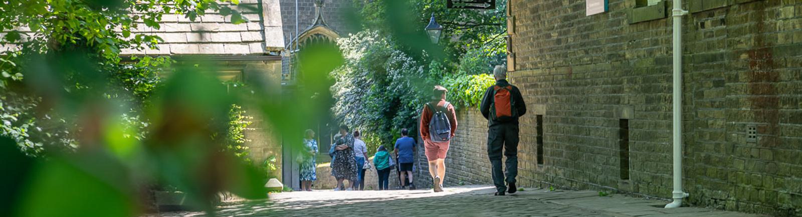 Photo of people walking down a cobbled street in dappled sunshine, with 19th-century buildings either side, framed by green trees.