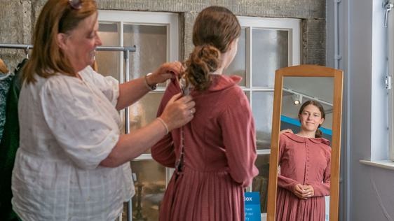 Photo of a lady helping a teenage girl into a Victorian-style costume in front of a mirror.