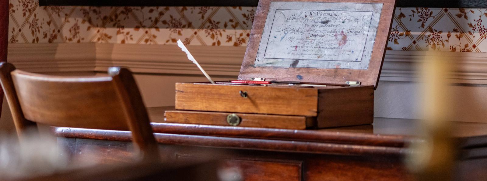Photo of a dark wooden chair and desk, with an old fashioned paint box sitting on top.