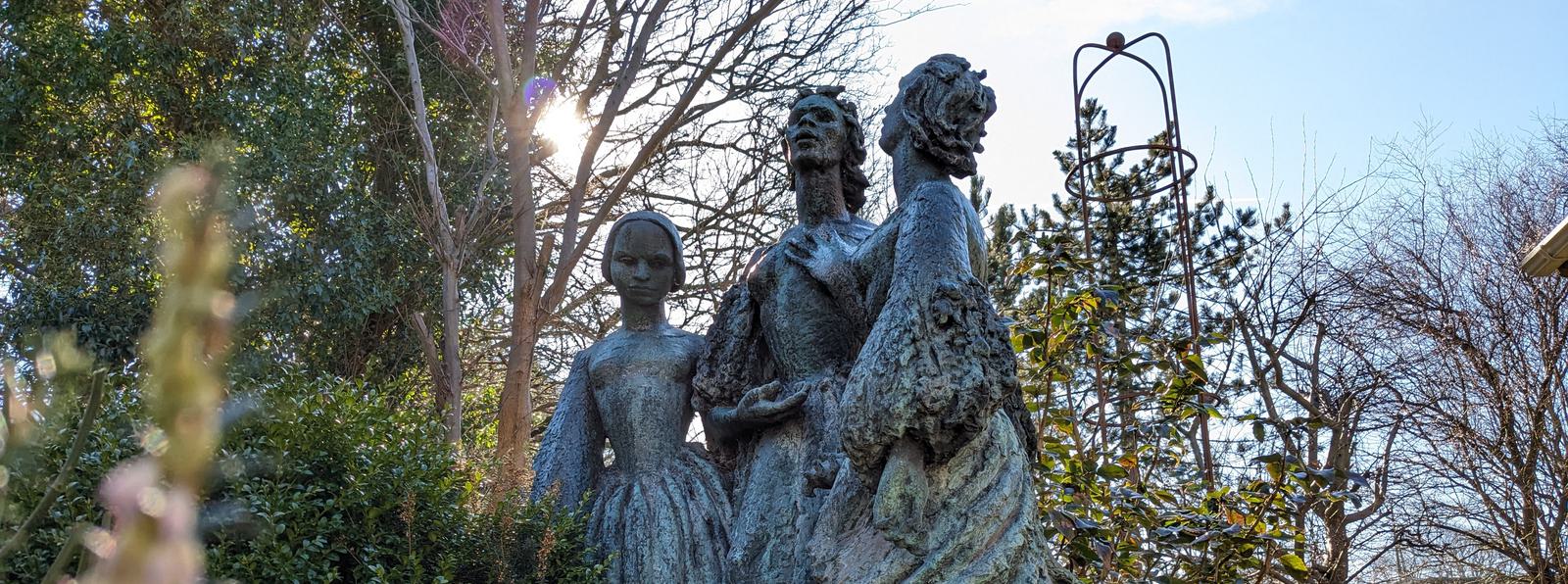 Photo of a statue of three women in early-19th-century clothing, in a garden.