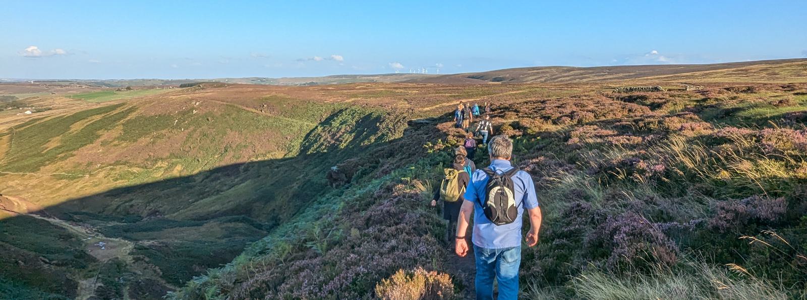 Photo of a line of people from behind, walking across sweeping moorland as the sun sets.