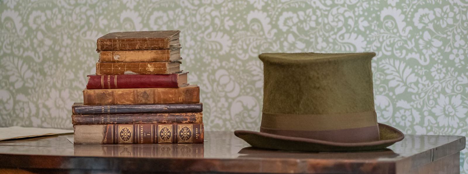 Photo of a pile of antique books on the left of a dresser and a soft green top hat on the right.