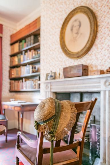 Photo of a wooden chair with a straw hat on it, in a room decorated in 19th-century style.